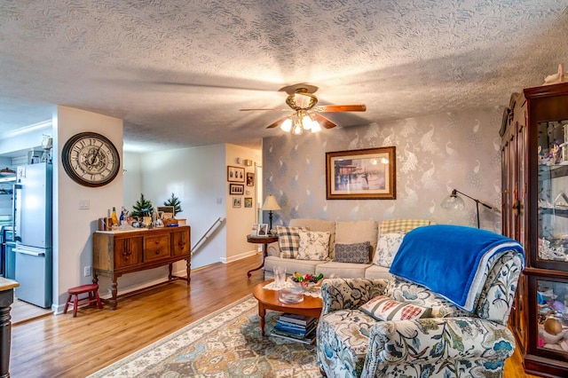 living room featuring a textured ceiling, wood finished floors, a ceiling fan, baseboards, and wallpapered walls