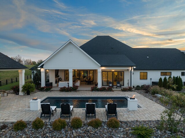 back of house at dusk featuring roof with shingles, fence, an outdoor pool, and a patio