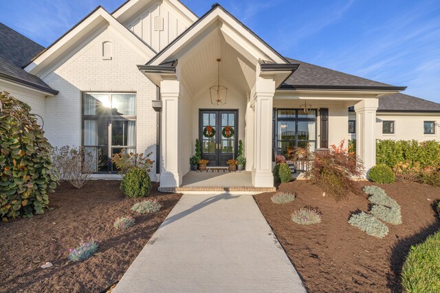 view of exterior entry featuring roof with shingles, covered porch, french doors, board and batten siding, and brick siding