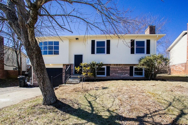 raised ranch with brick siding, concrete driveway, a chimney, an attached garage, and stucco siding