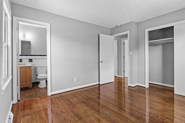 unfurnished bedroom featuring ensuite bath, wood finished floors, a closet, tile walls, and baseboards