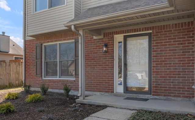 entrance to property with brick siding, a shingled roof, and fence