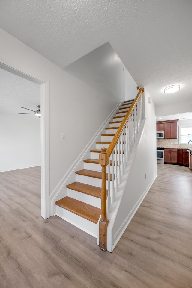 staircase with ceiling fan, wood finished floors, baseboards, and a textured ceiling