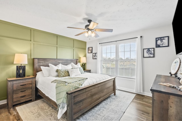 bedroom with a ceiling fan, a textured ceiling, wood finished floors, a decorative wall, and baseboards