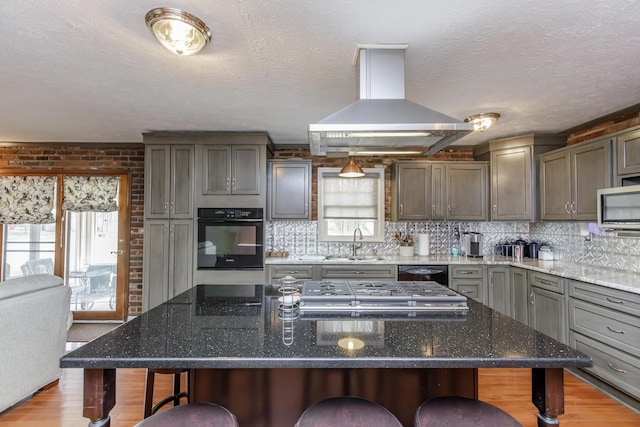 kitchen with black appliances, a sink, a breakfast bar area, island range hood, and decorative backsplash