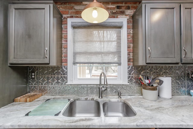 kitchen with gray cabinets, a sink, light stone counters, backsplash, and brick wall