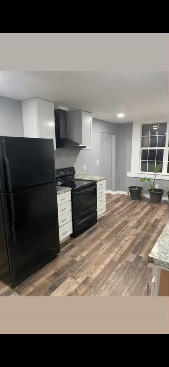 kitchen with wood finished floors, light stone countertops, black appliances, white cabinetry, and wall chimney exhaust hood