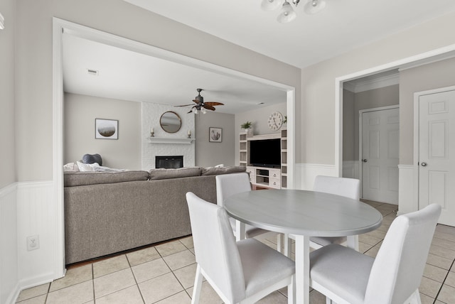 dining area with light tile patterned floors, a ceiling fan, a fireplace, and wainscoting