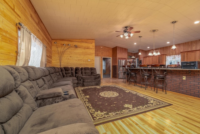 living room featuring vaulted ceiling, wooden walls, ceiling fan with notable chandelier, and light wood finished floors