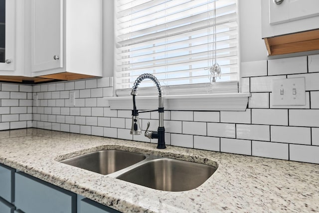 kitchen with a sink, light stone counters, backsplash, white cabinetry, and glass insert cabinets