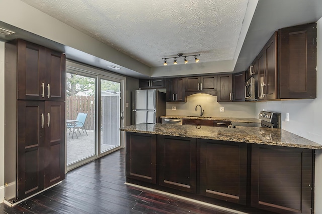 kitchen with dark stone countertops, appliances with stainless steel finishes, a peninsula, dark wood-style floors, and a sink