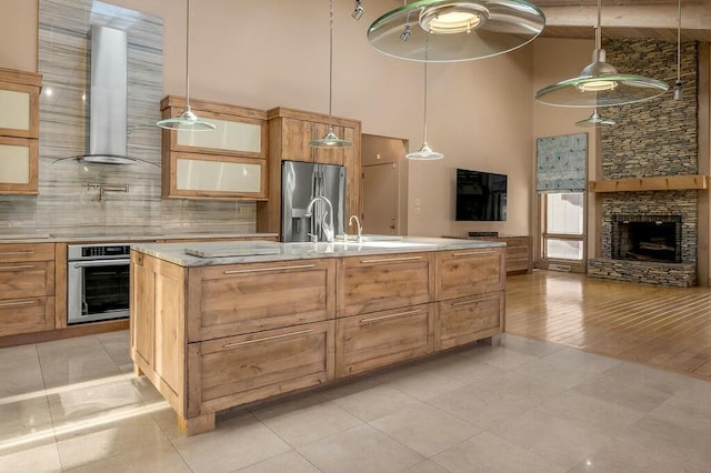 kitchen with a kitchen island with sink, stainless steel appliances, a stone fireplace, light countertops, and wall chimney exhaust hood