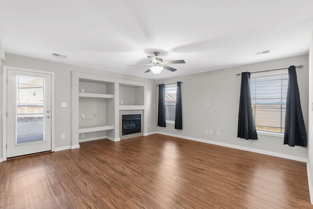 unfurnished living room featuring visible vents, built in shelves, a tiled fireplace, a healthy amount of sunlight, and dark wood-style flooring