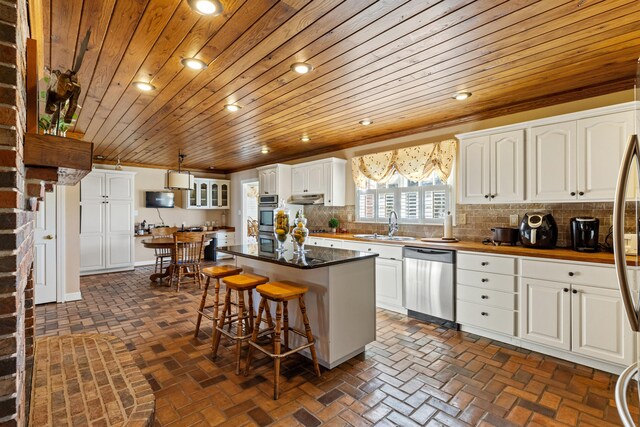 kitchen with brick floor, stainless steel appliances, wood ceiling, white cabinetry, and backsplash