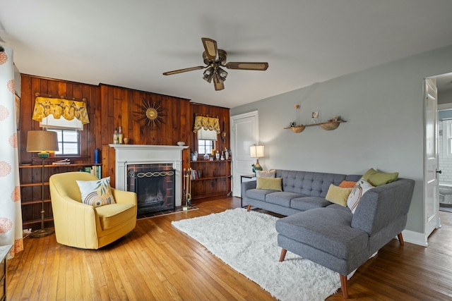 living room featuring a ceiling fan, a tiled fireplace, hardwood / wood-style flooring, wooden walls, and baseboards