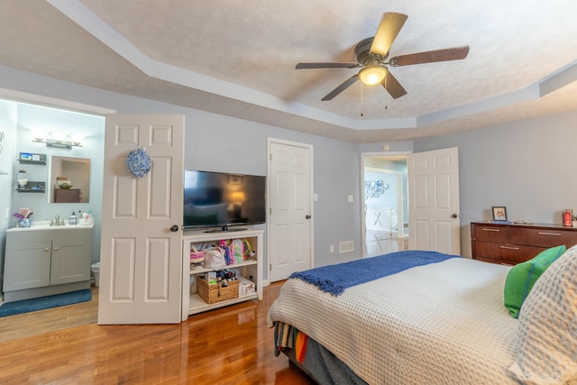 bedroom featuring connected bathroom, a textured ceiling, light wood-style floors, and a tray ceiling