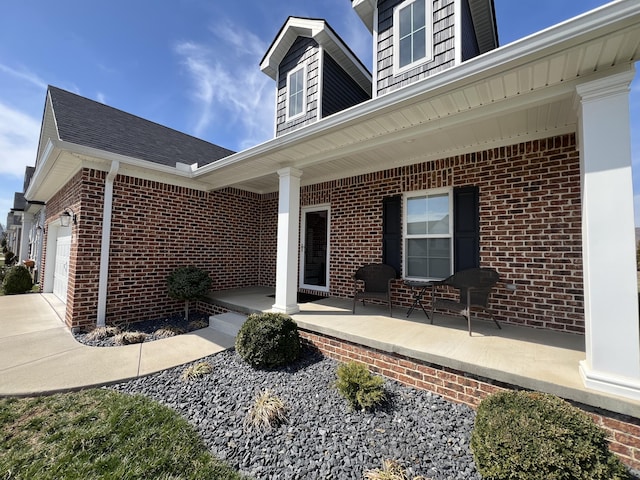 doorway to property featuring brick siding, a porch, an attached garage, and roof with shingles