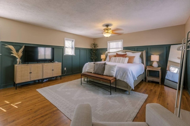 bedroom with a ceiling fan, a textured ceiling, wood finished floors, wainscoting, and a decorative wall