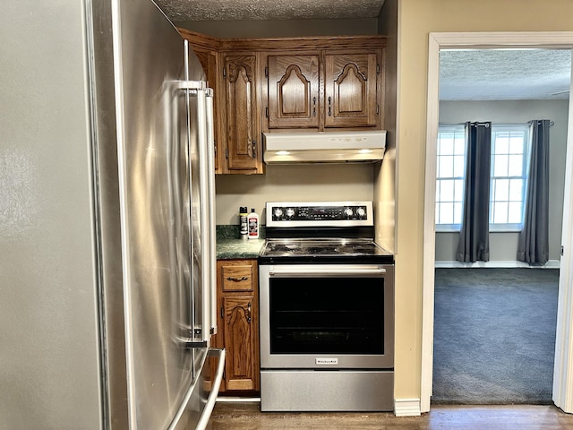 kitchen featuring dark countertops, under cabinet range hood, stainless steel appliances, a textured ceiling, and dark colored carpet