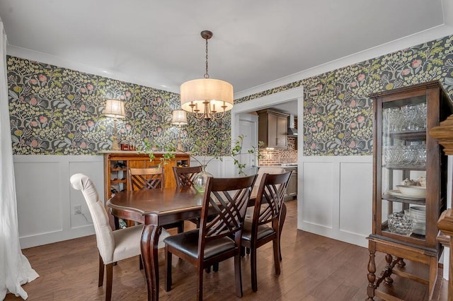 dining space featuring a wainscoted wall, wallpapered walls, dark wood finished floors, a decorative wall, and a chandelier