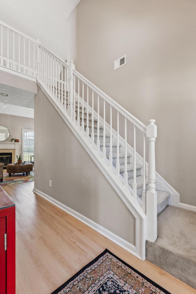 stairway featuring visible vents, baseboards, a towering ceiling, wood finished floors, and a glass covered fireplace