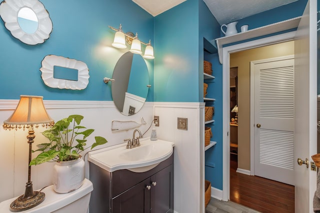 half bath with vanity, toilet, wood finished floors, and a wainscoted wall