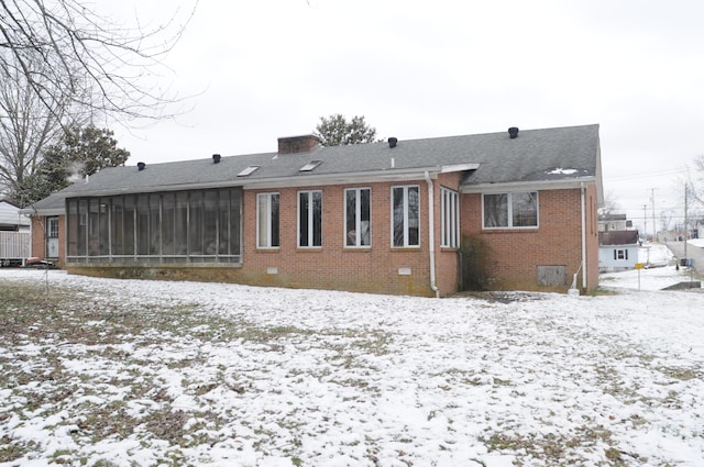 snow covered rear of property featuring roof with shingles, a sunroom, a chimney, crawl space, and brick siding