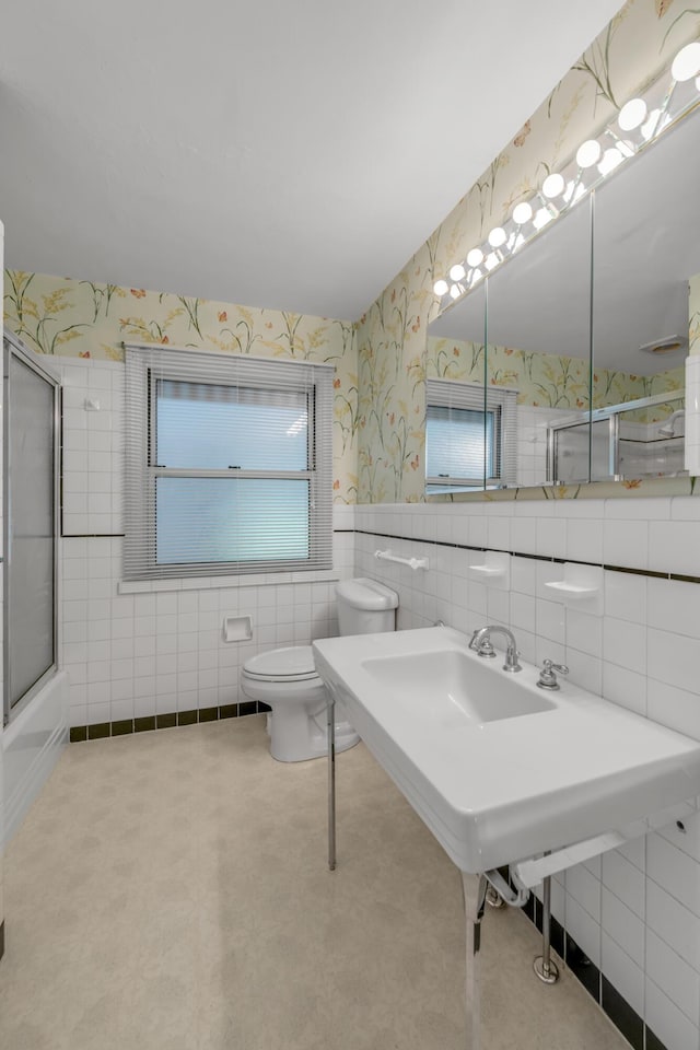 bathroom featuring a wainscoted wall, toilet, combined bath / shower with glass door, tile walls, and wallpapered walls