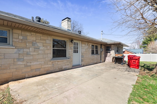 back of house with fence, a shingled roof, a chimney, stone siding, and a patio area