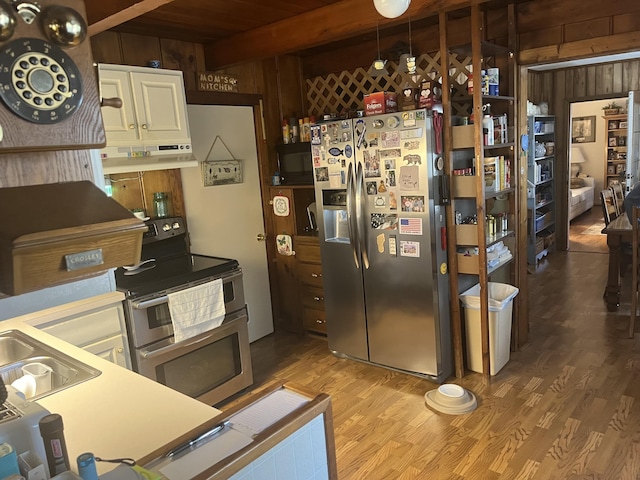 kitchen with wooden walls, stainless steel appliances, under cabinet range hood, beamed ceiling, and light wood-type flooring