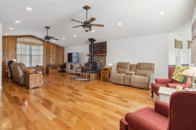 living area featuring light wood-style flooring, wooden walls, ceiling fan, a wood stove, and vaulted ceiling