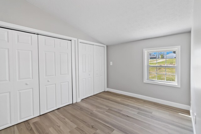 unfurnished bedroom featuring baseboards, light wood finished floors, vaulted ceiling, and two closets