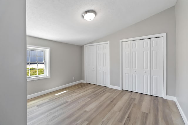 unfurnished bedroom with baseboards, two closets, a textured ceiling, vaulted ceiling, and light wood-style flooring