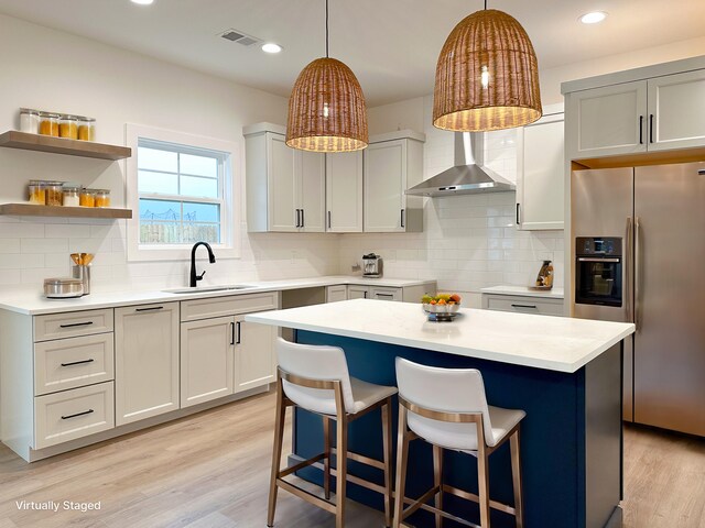 kitchen with a breakfast bar, a sink, stainless steel refrigerator with ice dispenser, light wood-style floors, and wall chimney range hood