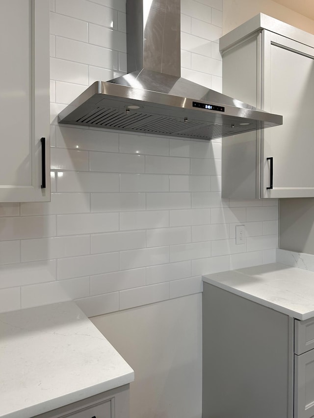 kitchen with wall chimney range hood, light stone countertops, backsplash, and gray cabinetry