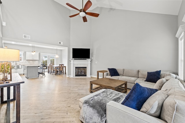 living room featuring light wood-type flooring, a fireplace with flush hearth, visible vents, ceiling fan, and high vaulted ceiling
