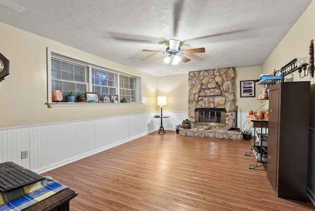 interior space featuring wainscoting, wood finished floors, a stone fireplace, ceiling fan, and a textured ceiling