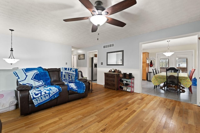 living area with a textured ceiling, a wainscoted wall, a ceiling fan, light wood-type flooring, and visible vents