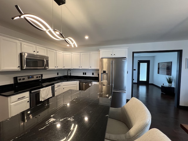 kitchen with white cabinets, hanging light fixtures, appliances with stainless steel finishes, a sink, and dark stone countertops
