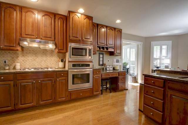 kitchen with appliances with stainless steel finishes, tasteful backsplash, brown cabinets, light wood finished floors, and under cabinet range hood