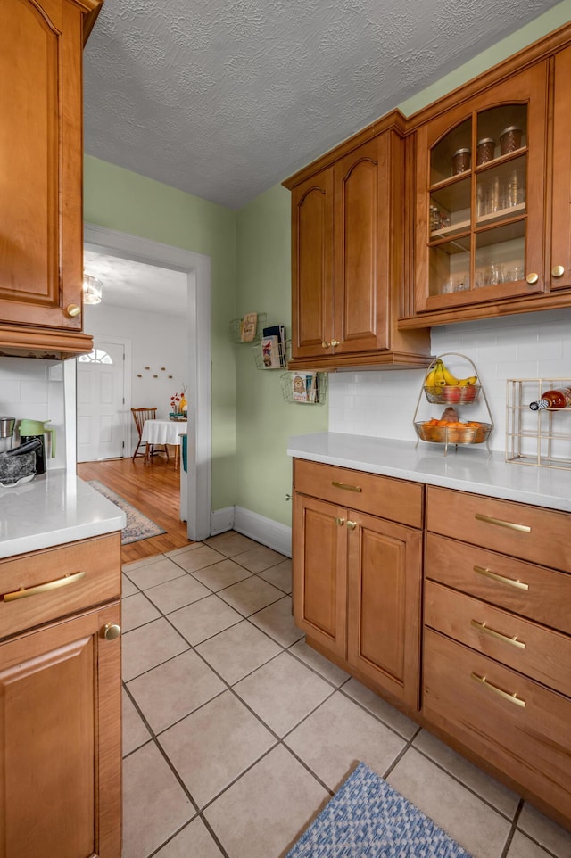 kitchen with light countertops, brown cabinetry, light tile patterned floors, glass insert cabinets, and tasteful backsplash