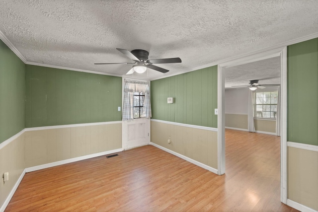 empty room with a textured ceiling, a ceiling fan, wood finished floors, visible vents, and ornamental molding