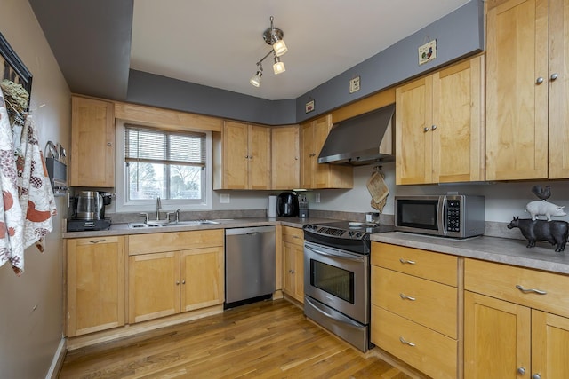 kitchen with light brown cabinetry, a sink, light wood-type flooring, wall chimney exhaust hood, and appliances with stainless steel finishes