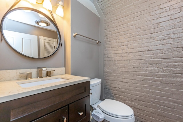 bathroom featuring toilet, vanity, and brick wall