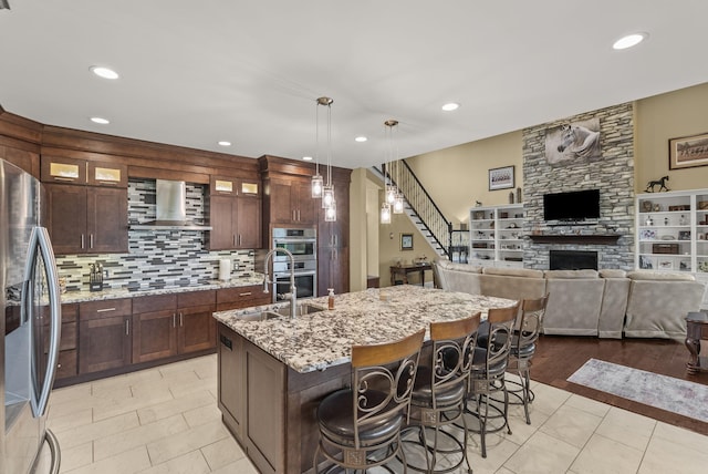 kitchen featuring stainless steel appliances, wall chimney exhaust hood, a stone fireplace, open floor plan, and a center island with sink