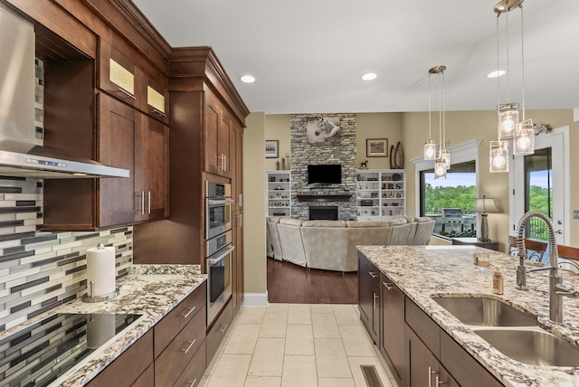 kitchen featuring a sink, black electric cooktop, wall chimney exhaust hood, a stone fireplace, and open floor plan