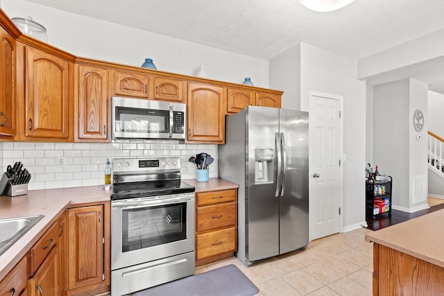 kitchen with light tile patterned floors, stainless steel appliances, brown cabinetry, and decorative backsplash