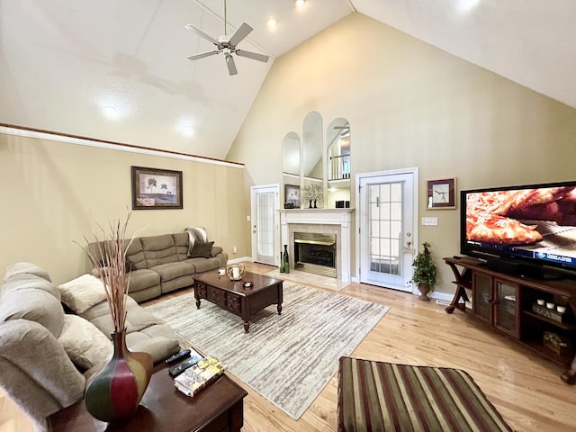 living area featuring ceiling fan, baseboards, light wood-type flooring, a fireplace, and high vaulted ceiling