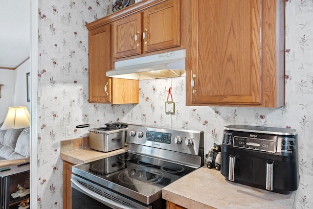 kitchen with electric stove, under cabinet range hood, brown cabinetry, light countertops, and wallpapered walls