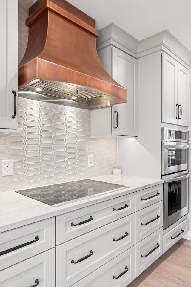 kitchen featuring decorative backsplash, black electric cooktop, premium range hood, white cabinets, and double oven